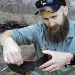 Mężczyzna w czapce z daszkiem z obfitą brodą, jest na wykopaliskach. Trzyma fragmenty ceramiki